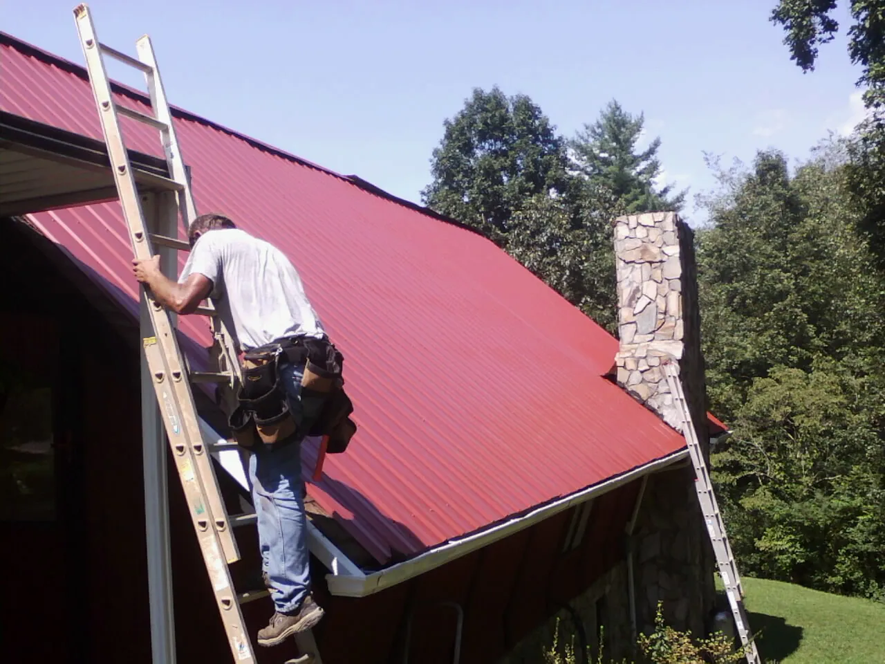 Roofer on a ladder beside a red metal roof and stone chimney — Soffit & Fascia Repair by Roofing Brothers in Sullivan